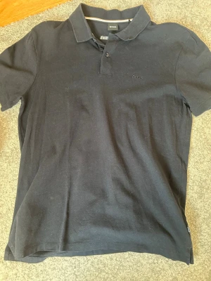 Svart pikétröja från BOSS - Klassisk svart pikétröja från BOSS med diskret broderad logga på bröstet. Tröjan har korta ärmar, krage och knappar vid halsen. Tillverkad i mjuk bomull som känns skön mot huden. Perfekt för en clean och stilren look.