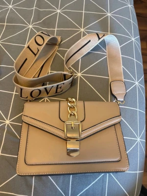 Beige axelväska med bred rem - Snygg beige axelväska med bred textilrem där det står 'LOVE' i svart. Väskan har guldfärgade detaljer, kedja och spänne framtill. Tillverkad i slätt konstläder och har en stilren, rektangulär form. Perfekt för dig som vill ha en trendig accessoar.