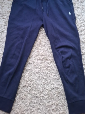 Mörkblå mjukisbyxor från Polo Ralph Lauren - Säljer ett par mörkblå mjukisbyxor från Polo Ralph Lauren med klassisk vit logga broderad på vänster sida. Byxorna har dragsko i midjan och muddar vid bensluten. Perfekta för chill dagar och riktigt sköna tack vare mjukt material.