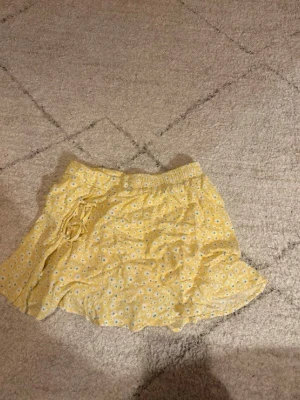 Gula blommig kjol med snörning - Söta gula kjol med små vita blommor och snörning i midjan. De har en avslappnad passform och elastisk midja för extra komfort. Perfekta för varma dagar och ger en fräsch sommarkänsla.