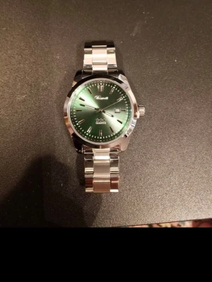 Klassisk silverfärgad klocka med grön urtavla - Stilren klocka från Kiseki med metallarmband i silver och en grön urtavla. Klockan har tydliga markeringar, datumvisning och klassisk rund form. Perfekt accessoar för dig som gillar en tidlös look.