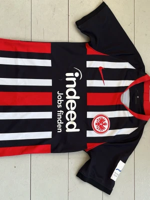 Eintracht Frankfurt matchtröja Nike - Snygg Eintracht Frankfurt matchtröja från Nike med svart, rött och vitt randigt mönster. Tröjan har klubbmärke på bröstet och röd V-ringning Perfekt för fotbollsfans som vill sticka ut på planen.