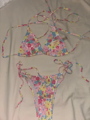 Färgglad blommig bikini med knyt - Säljer en superfin trekantsbikini med blommigt mönster i rosa, blått, gult och grönt. Bikinin har smockad struktur och knyt både i sidan på underdelen och i nacken och ryggen på överdelen. Perfekt för sommaren och strandhäng!