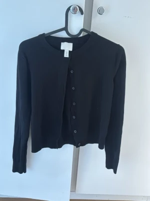 Svart basic kofta från H&M - En enkel svart kofta från H&M med rund halsringning och knäppning framtill. Koftan har långa ärmar och en rak passform, perfekt att slänga över en topp eller klänning. Tillverkad i mjukt och stretchigt material.