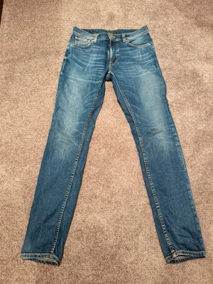 Blå slim jeans från Nudie Jeans - Säljer ett par klassiska blå slim fit jeans från Nudie Jeans. Jeansen har fem fickor, snygga slitningar och kontrastsömmar. Perfekta till en avslappnad stil och tillverkade i mjuk bomull för skön känsla hela dagen.