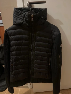 Svart Cardigan från Moncler - Säljer en svart pufferjacka från Moncler med quiltad front och rygg, stickade ärmar och huva. Jackan har dragkedja framtill, två fickor med knapp och en liten ficka med dragkedja på ärmen. Perfekt för kalla vinterdagar och riktigt snygg streetstil.