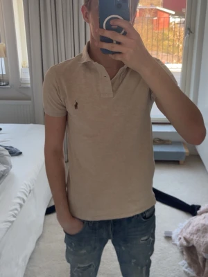 Beige pikétröja från Polo Ralph Lauren - Klassisk beige pikétröja från Polo Ralph Lauren med korta ärmar och krage. Tröjan har två knappar vid halsen och den ikoniska broderade loggan på bröstet. Tillverkad i mjuk bomull för en bekväm känsla och snygg passform.