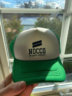 Grön och vit NOCCO truckerkeps - Säljer en grön och vit truckerkeps från NOCCO med klassisk mesh på sidorna och justerbar snapback. Framsidan har NOCCO-logga och texten 'No Carbs Company' i svart. Perfekt för dig som gillar sportig och avslappnad stil. En liten fläck som inte syns på bild men kan visas!