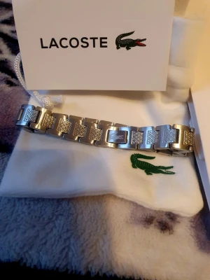 Silverfärgat armband från Lacoste - Snyggt armband i silverfärgad metall från Lacoste med länkdesign och ingraverade krokodil-loggor på varje länk. Armbandet har en modern och stilren look, perfekt för dig som gillar accessoarer med en sportig touch. Levereras med originalpåse från Lacoste.