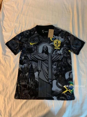 Svart Brasilien Nike fotbollströja - Unik svart fotbollströja från Nike med Brasilien-emblem och gult Nike-märke. Tröjan har ett stort tryck av Kristusstatyn och detaljerade grafiska mönster i grått och svart. Rund hals och korta ärmar. Perfekt för dig som vill sticka ut på planen.