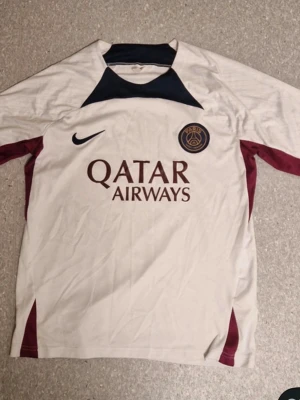 PSG matchtröja vit Nike - Snygg vit PSG matchtröja från Nike med Qatar Airways-tryck på bröstet och klubbmärke. Marinblå detalj vid halsen och vinröda inslag på sidorna. Perfekt för fotbollsträning eller att bära till vardags. Tillverkad i lätt och ventilerande material.