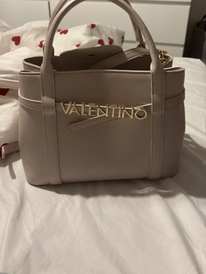 Ljusbeige handväska från Valentino - Säljer en ljusbeige handväska från Valentino med guldfärgad logga framtill. Väskan har två handtag, avtagbar axelrem och stilren, fyrkantig form. Tillverkad i slätt konstläder och stängs med magnetknapp. Perfekt för dig som gillar klassisk och trendig design.