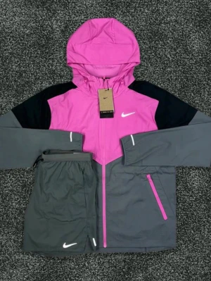 Nike träningsset rosa/grå - Snyggt träningsset från Nike med jacka och byxor. Jackan är rosa och grå med svarta axelpartier, huva och dragkedja framtill. Byxorna är grå med vit Nike-logga och reflexdetaljer. Perfekt för löpning eller utomhusträning.