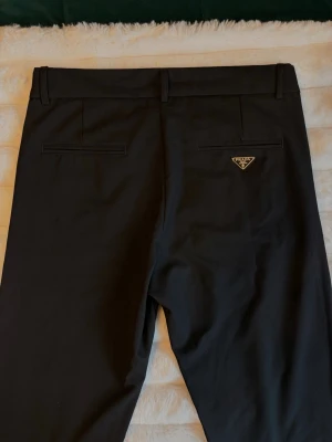 Kostymbyxor från Prada - Säljer ett par äkta Prada-byxor i svart. Modell som passar perfekt både till vardag och mer uppklädda tillfällen.  Byxorna är i mycket fint skick och håller hög kvalitet, använda 2 gånger men ser helt nya ut.  Storlek: 48 Färg: Svart Material: 100% bomull Nypris: 14.000kr  Observera att kvitto saknas, men äktheten kan enkelt verifieras via QR-koden och märkningen på insidan (se bilder). Alla etiketter finns kvar.
