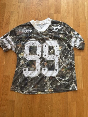Camouflage mesh t-shirt med nummer 99 - Säljer en sportig t-shirt i meshmaterial med camouflage-mönster och stort vitt nummer 99 framtill. T-shirten har korta ärmar, vit v-ringad krage och stjärndetaljer. Perfekt för dig som gillar streetwear och vill sticka ut.