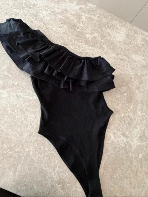 Eftertraktad Zara body med volanger - Snygg svart body med one shoulder-design och flera lager volanger över axeln. Tight passform och högt skuren vid benen. Perfekt för dig som vill sticka ut med en trendig och stilren look. Materialet känns mjukt och stretchigt.