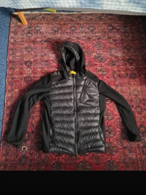 Svart Parajumper - Säljer en svart pufferjacka med huva och dragkedja. Jackan har quiltade partier fram och bak, släta ärmar och sidopaneler i mjukare material. Bröstficka med dragkedja och två sidofickor. Perfekt för kalla dagar och riktigt snygg streetstil.