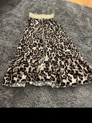 Leopardmönstrad långkjol med snörning - Snygg långkjol från New Collection med leopardmönster och bred, elastisk midja med snörning. Kjolen har en flowy passform och är perfekt för dig som vill sticka ut med ett djurmönster. Färgerna går i beige, svart och brunt.