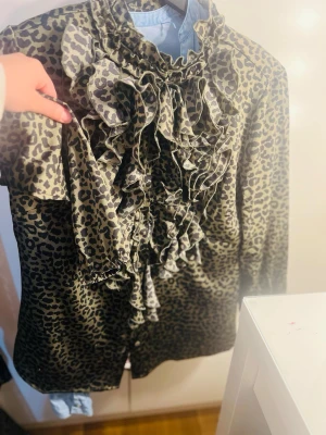 Leopardmönstrad blus med volanger - Snygg blus med leopardmönster och volangdetaljer framtill. Blusen har hög krage och långa ärmar, perfekt för dig som vill sticka ut med en trendig look. Materialet känns mjukt och följsamt mot huden.
