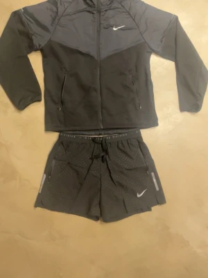Nike träningsset jacka & shorts - Snyggt träningsset från Nike med svart/grå långärmad jacka med huva och dragkedja samt matchande svarta shorts. Jackan har reflexdetaljer och Nike-logga både fram och bak. Shortsen har resår i midjan och små prickdetaljer. Perfekt för löpning eller gym.
