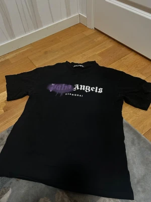 Palm Angels svart t-shirt Shanghai - Säljer en svart t-shirt från Palm Angels med trycket 'Palm Angels Shanghai' på bröstet. Texten har en cool lila spray-effekt över 'Palm' och resten av texten är vit. Klassisk passform och rund hals. Perfekt för dig som gillar streetwear.