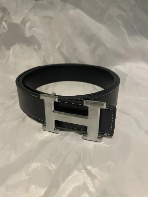 Svart skärp med silver H-spänne - Stilrent svart bälte med ett stort H-format spänne i silverfärgad metall. Bältet har vita sömmar längs kanterna och är tillverkat i slätt skinn. Perfekt accessoar för att lyfta din outfit.110cm