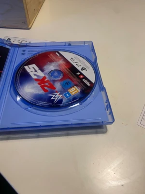 WWE 2K23 PS5 spel - WWE 2K23 till PlayStation 5, ett actionfyllt wrestling-spel med snyggt omslag i blått, rött och vitt. Skivan är i klassisk rund form och levereras i ett blått plastfodral. Perfekt för dig som gillar sport och fighting på konsol.