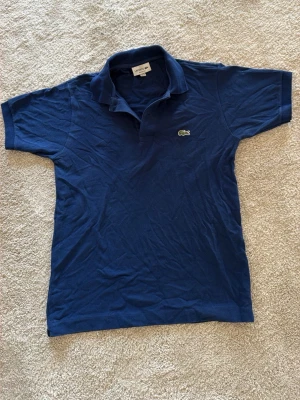 Blå pikétopp från Lacoste - Snygg blå pikétopp från Lacoste med klassisk krage och knappar vid halsen. Den har korta ärmar och den ikoniska gröna krokodilloggan broderad på bröstet. Tillverkad i mjuk bomull som känns skön mot huden. Pris kan diskuteras