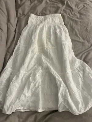 Vit broderad långkjol med resår - Säljer en vit långkjol med broderade blommor och resår i midjan. Kjolen har en luftig och flowy passform med fina hålbroderier som ger en romantisk känsla. Perfekt till sommaren och lätt att matcha med olika toppar.