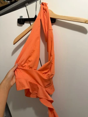 Orange omlottopp med volang - Supersnygg orange omlottopp med knyt i nacken och volangdetalj nedtill. Toppen är ärmlös och croppad, perfekt för dig som gillar färg och trendiga detaljer. Materialet känns mjukt och följsamt mot huden.