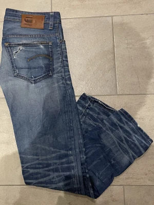 Blå jeans från G-Star RAW - Säljer ett par blå jeans från G-Star RAW med slitningar och snygg tvätt. Klassisk femficksmodell med raka ben och läderpatch bak i midjan. Jeansen har kontrastsömmar och detaljerade bakfickor. Perfekta för dig som gillar en avslappnad men ändå stilren look. 32/32