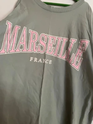 Grön oversized t-shirt MARSEILLE från Divided - Snygg grön t-shirt från Divided med stort rosa och vitt MARSEILLE-tryck på bröstet. Klassisk rund halsringning och normal passform. Perfekt för dig som gillar streetstyle och vill ha en chill vibe.