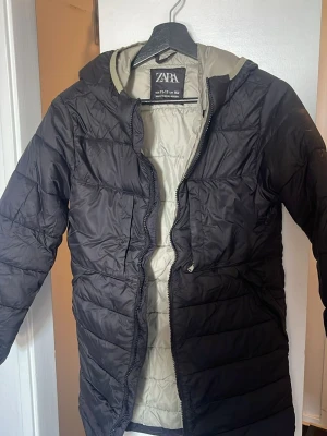 Svart pufferjacka från Zara - Säljer en svart pufferjacka från Zara med huva och dragkedja framtill. Jackan har quiltad design och ljus insida, perfekt för kalla vinterdagar. Enkel och stilren modell som håller dig varm och bekväm.