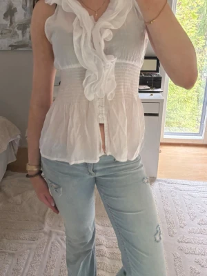 Vit volangblus från TSEBO - Supersöt vit blus från TSEBO med volangdetaljer längs knäppningen framtill. Ärmlös modell med smock i midjan som ger en snygg passform. Perfekt att styla med jeans eller kjol för en fräsch look.