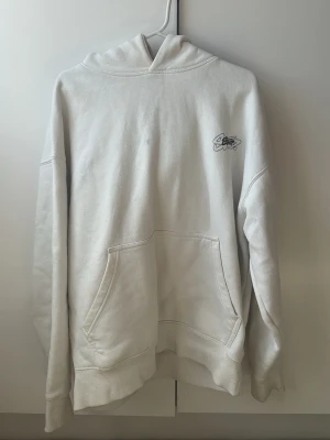 Vit hoodie Street Style - Vit hoodie med coolt street style-tryck på ryggen och mindre logga på bröstet. Klassisk passform med huva och stor magficka. Materialet är mjukt och bekvämt, perfekt för dig som gillar en avslappnad look och grafiska detaljer.