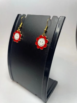 Röda blommiga örhängen med smiley - Säljer ett par söta örhängen i form av röda blommor med vit smiley i mitten. Örhängena har guldfärgade krokar och är tillverkade i metall och emalj. Perfekta för dig som vill ha en lekfull och färgglad accessoar till din stil.