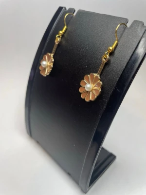 Blommiga örhängen med pärldetalj - Snygga örhängen i guldton med blomformad design och en liten vit pärla i mitten. Blommorna har ljusrosa emaljblad och örhängena har krokfäste i metall. Perfekta för dig som gillar söta och unika accessoarer.