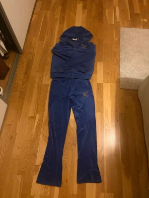 Blå velour loungewear-set JC - Mysigt blått loungewear-set från Juicy Couture i mjuk velour. Setet består av en zip-hoodie med huva och matchande byxor med strassdetaljer och JC-logga på benet. Perfekt för chill dagar hemma.