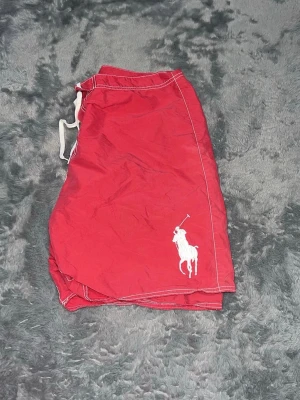 Polo Ralph Lauren Big logo shorts - Riktigt feta röda badshorts från Polo Ralph Lauren med den ikoniska "Big Pony"-loggan broderad i vitt. Snygga detaljer med R3 på sidan och vita kontrastsömmar. Perfekta till stranden eller som sköna sommarshorts. • Passform: Klassisk modell med dragsko i midjan så man kan justera passformen. Sitter lagom baggy och skönt. • Detaljer: Stor broderad logga, sidficka med knäppning och bakficka. • Skick: Toppskick, nytvättade och redo för sommaren! 