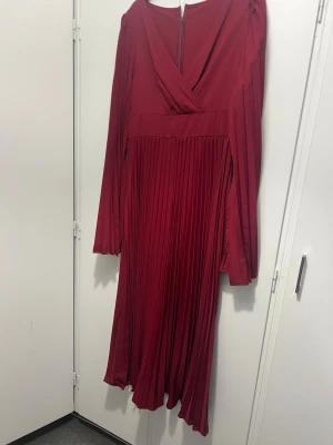 Vinröd plisserad långklänning - Snygg vinröd långklänning med plisserad kjol och långa ärmar. Klänningen har omlott upptill och vacker v-ringning. Tillverkad i ett glansigt material som ger en elegant look. Perfekt för dig som vill sticka ut med stilrena detaljer.