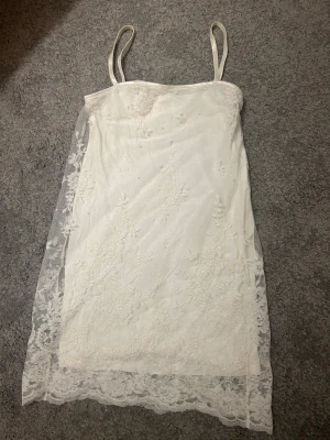 Vit spetsklänning från Ida Sjöstedt - vit klänning från Ida Sjöstedt med tunna axelband och vacker spetsdetalj över hela plagget. Klänningen har en rak passform och är täckt med blommig spets. Perfekt för sommarens alla tillfällen.