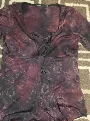 Mörklila topp med snörning - Unik mörklila topp med blommigt mönster och snygg snörning framtill. Toppen har en djup, fyrkantig ringning och är tillverkad i ett mjukt, stretchigt material. Perfekt för dig som vill sticka ut med coola detaljer och boho/whimsical-vibe.