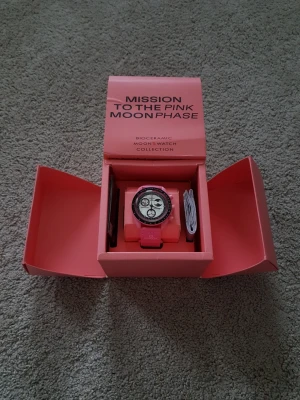 Omega x Swatch Pink Moonphase klocka - Säljer en Omega x Swatch MoonSwatch Mission to the Pink Moonphase. Klockan har en rosa boett och armband, vit urtavla med svarta detaljer och månfaskomplikation. Kommer i originalförpackning. Perfekt för dig som gillar unika accessoarer!