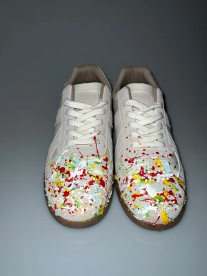 Maison margielas med färgstänk  - Unika vita sneakers med färgstänk i rött, gult och grönt på tån. Skorna har klassisk låg siluett, vita snören och bruna gummisulor. Tillverkade i läder och mocka för en stilren men kreativ look.                                                                         Till behör- du får med boxen                                                                                                 Och ett par crease skydd! 