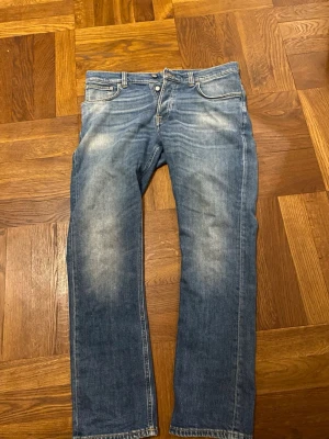 Nudie jeans - Säljer ett par klassiska blå jeans med raka ben och snygga slitningar framtill. Jeansen har fem fickor, normal midja och kontrastsömmar. Perfekta för en avslappnad och trendig look. Materialet är denim i bomull. Modell: Grim Tim. Storlek: W36 L32