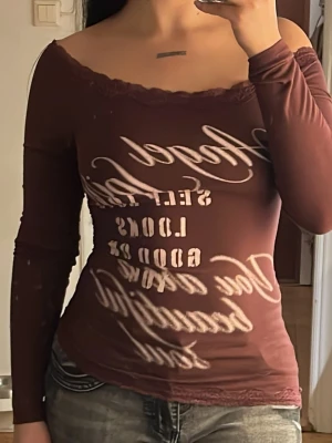 Brun offshoulder topp med text - Säljer en brun offshoulder topp med lång ärm och dekorativ spetskant vid halsringningen. Toppen har ett tryckt textmotiv framtill och är figurnära i passformen. Perfekt för dig som gillar en trendig och unik stil.
