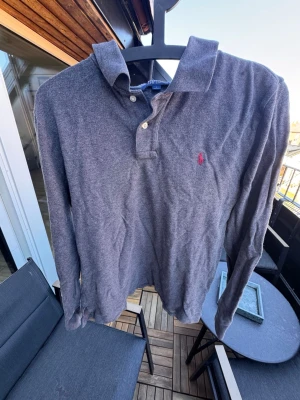 Mörkgrå långärmad  från Polo Ralph Lauren🙌🤩 - Säljer en mörkgrå långärmad henley från Polo Ralph Lauren med klassisk krage och två knappar vid halsen. Skjortan har det ikoniska röda Polo-logotypen broderad på bröstet och är tillverkad i mjuk bomull. Perfekt för en avslappnad men ändå stilren look.🙌🤩