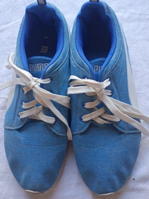 Blå sneakers från Puma med vita detaljer - Snygga blå sneakers från Puma med vita sidor och vita skosnören. Skorna har en låg profil och är tillverkade i textilmaterial med blå innerfoder. Perfekta för dig som gillar sportig och avslappnad stil.