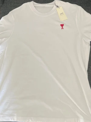 Vit t-shirt från Ami med rött hjärta - Säljer en clean vit t-shirt från Ami Alexandre Mattiussi med ett broderat rött hjärta och bokstaven A på bröstet. Klassisk rund hals och mjuk bomullskvalitet. Perfekt för dig som gillar stilrena plagg med en twist.
