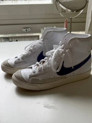Nike vita höga sneakers  - Säljer ett par klassiska Nike sneakers i vitt med blå swoosh på sidan. Skorna är höga och har vita snören, tåparti i mocka och ovandel i läder. 